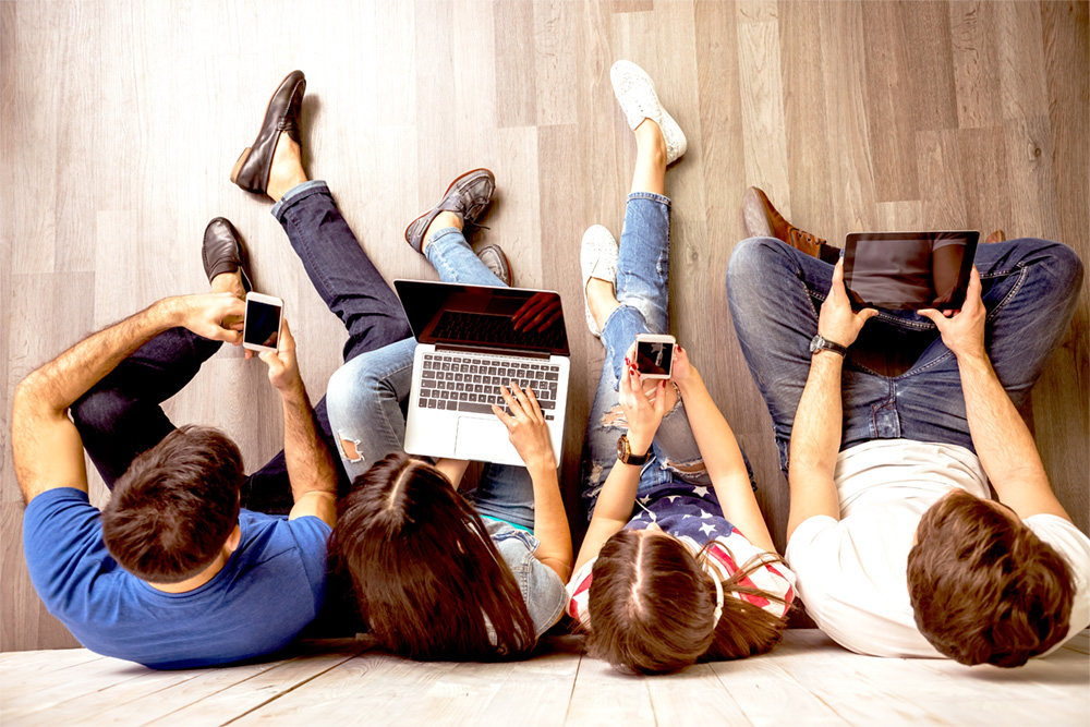 Young people sitting on floor against wall looking at electronic devices